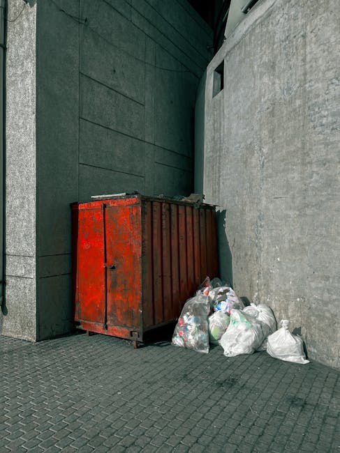 A worker wearing a yellow high-visibility vest and dark trousers is in front of a large red waste collection vehicle on a street. The worker is positioned near the rear of the truck, which has an open