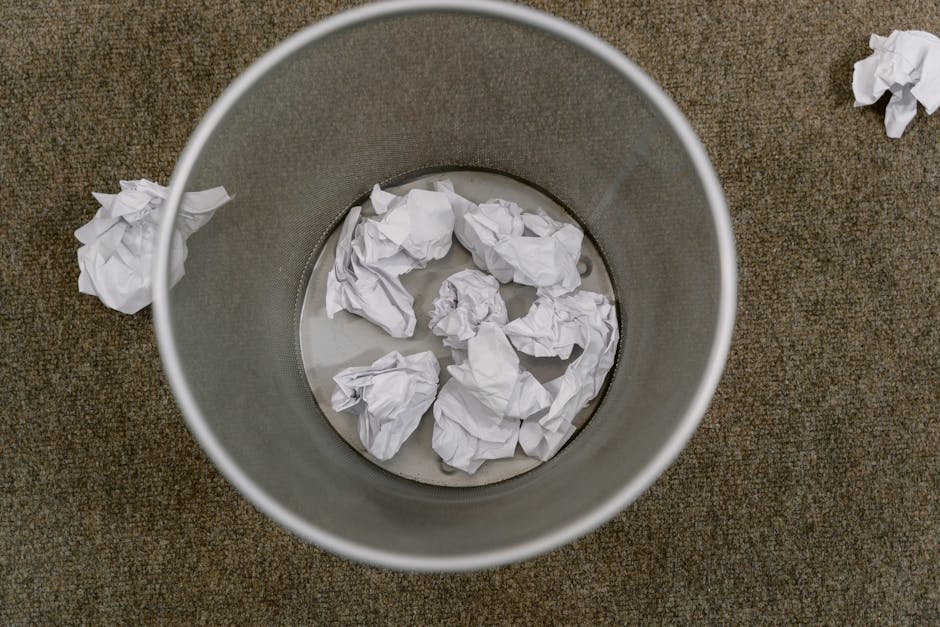 A cylindrical waste bin made of grey metal mesh is filled with crumpled white paper, positioned on a carpeted floor. Several loose crumpled papers are scattered on the beige-brown carpet nearby, with
