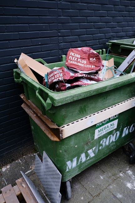 A large pile of mixed household waste, including black and clear plastic rubbish bags, flatten cardboard boxes, paper and packaging materials, and a few unopened parcels, situated on an urban paved pa