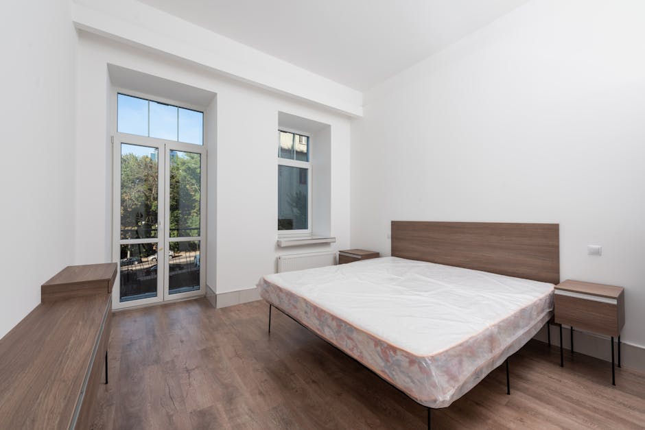 A minimalistic, modern bedroom with white walls and a white ceiling, featuring a large bed with a plain white mattress and a padded, vertical-serrated white headboard positioned against the right wall